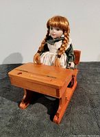 Front view of Anne of Green Gables porcelain doll seated at wooden school desk, showing detailed doll features and desk top.