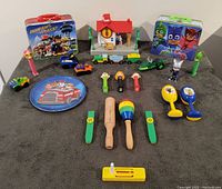 Assortment of children's toys including Paw Patrol themed lunchbox, plate, small toy vehicles, PJ Masks lunchbox, Pez dispenser, Melissa & Doug musical instruments, and Little People train station.