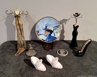 Full view of assorted jewelry holders, shoe ring holder, Avon collector plate on stand, ceramic baby shoes, small glass ring holder, and round compact mirror.