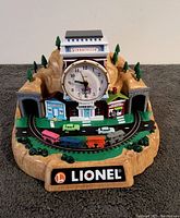 Front view of Lionel 100th Anniversary alarm clock designed as a mountain landscape with clock face, track decoration, buildings, trees, and small vintage metal train cars.