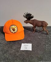 Orange hunting cap with Moose and Deere Hunter Ontario embroidered patch next to a large resin moose figure standing on all fours with prominent antlers.