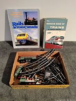 Image of a cardboard box containing two vintage train books and a partial toy train set with tracks and power cord.