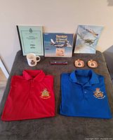 Overview photo showing all lot items including shirts, books, mug, pen, ashtrays, and poster.