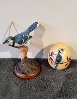 Photo showing the Blue Jay wood carving on branch stand next to the painted wooden egg with Blue Jay design, on a grey cloth background, against a white wall.