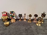 Nine assorted vintage brass bells arranged in a row on gray fabric against a white background showing variety in size and style.