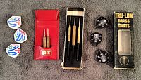 Top view showing three white/red/blue flights on the left, red leather M-Y branded carrier holding two darts with wooden tips, three brass darts with black shaft tips in original Tru-Lon box in the center, and three black skull and crossbones flights on the right.