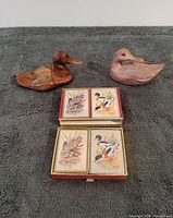 Two hand-carved wooden duck figurines placed next to a double deck of sealed playing cards featuring duck artwork.