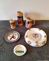 Lot overview showing three ceramic German beer steins, one large porcelain ashtray with German heraldry, and two smaller ashtrays (one white porcelain branded Morosoff Vodka, one pewter with illustration)