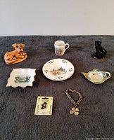 All items from the vintage Irish souvenir lot displayed on a grey fabric background including stoneware dog figurine, black ceramic cat figurine, pottery dishes, pin, and bracelet.