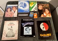 All vintage music and instrument items arranged on a dark surface: musical souvenir brochures for The King and I, Cabaret; instrument guides including Hohner booklet; Superman The Movie sheet music; and Hero harmonica.