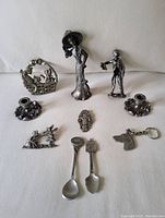Full view of all assorted vintage pewter items including figurines, spoons, pin, keychain, and candle holders arranged on white background.