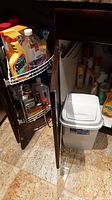 Contents of the under sink cabinet showing cleaning products on wire shelves and white plastic garbage pail