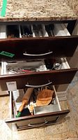Three kitchen drawers containing cutlery and utensils. Top drawer has organized silver-tone cutlery, middle drawer has various utensils including zester, jar opener, can opener, and bottom drawer contains wooden rolling pin, cutting board, and other utensils.