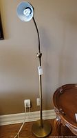 Full view of the brass-colored floor lamp with goose neck arm and white interior reflector on lamp head, standing beside a wooden table.