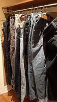 Side view of assorted men's pants and jeans hanging on a wooden rack, showing different colors and styles including denim and casual fabric pants.