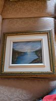 Framed original landscape painting by Gigi placed on a beige patterned surface, showing full framing and matting.