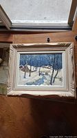 Framed original winter landscape painting showing snow-covered trees, a log cabin with smoke from chimney, and a creek.