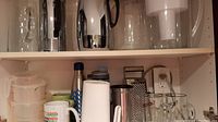 Top shelf cabinet showing two large jars, several pitchers and a water filter pitcher.