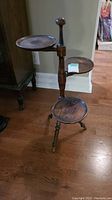 Full view of vintage wooden plant stand with three round shelves and tripod metal legs on hardwood floor.