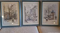Three framed and matted Paris street scene prints laid on a couch, showing each framed image in grey-blue frames with mats.