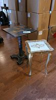 Two vintage occasional tables displayed together on hardwood floor in front of stacked cardboard boxes. One black wooden square top table with pedestal base and the other ornate gold and white decorative table with curved legs.