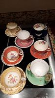 Top-down view showing all seven tea cups and saucers arranged closely together, each uniquely colored with floral patterns and gold trim.