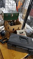 Two toolboxes visible with assorted tools, including a green metal toolbox, a cardboard box with tools inside, and a black plastic toolbox on a wooden surface.