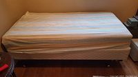 Twin bed made up with mattress, box spring and fitted striped sheet, on wooden frame with wheels visible.