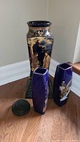 Three pottery vases on hardwood floor against wall: one tall black rectangular vase with Asian figures and gold detailing, two cobalt blue rectangular vases with floral and bird designs.