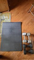 Photo of folding black exercise mat beside two pairs of dumbbells on wood floor.