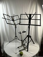 Three music stands together including two sheet music stands and one instrument stand with a green apple for size reference.