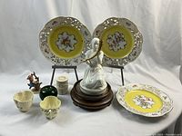 Full view of collection showing 3 Dresden plates with floral patterns and pierced rims, M.I. Hummel Apple Tree Girl figurine on wooden base, carved case with lighter, 2 Beleek teacups, a small green glass object, and Royal Copenhagen figurine.