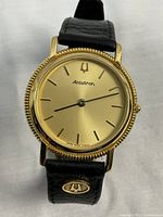 Front view of watch showing champagne dial, gold-tone case, black leather strap with Accutron logo.