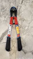 Front view of bolt cutters showing red handles with black grips and black cutting head, on marble surface.
