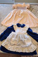 View of two girls' dresses, one light-colored with smocking, one navy with white apron