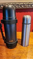 Large black Thermos flask and smaller stainless steel Thermos flask standing side by side