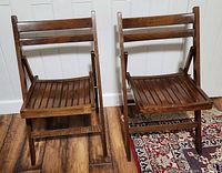 Front view of two dark stained solid wood folding chairs with slatted seats and backs on wooden floor and patterned rug.