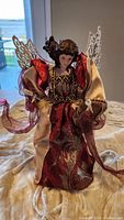 Angel tree topper displayed standing on cream fabric background, showing layered red and gold dress and gold wings.