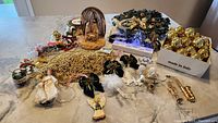 Photo displaying a variety of Christmas decorations spread on a table including a glass nativity scene with stable, gold bead garlands, assorted gold spherical ornaments, angel and ballerina figurines, bows, horses, and a holiday wreath.