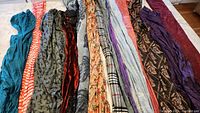 Lot of 13 scarves laid out in a row showing various colors, textures and patterns, including teal, orange stripes, floral prints and velvet.