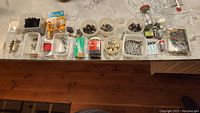 Wide view showing multiple containers filled with various screws, nails, picture hangers, L brackets, knobs, and small hardware items arranged on a marble countertop.