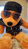 Front view of the plush bear wearing black leather vest, brown goggles, and black cap.