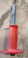 Top down view of the metal chisel with large red plastic guard placed on a marble surface, showing rust on the blade.