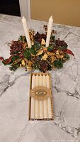 Christmas table centerpiece with two white tapered candles inserted, artificial greenery, red berries, and gold floral accents on a marble surface with a boxed set of six new white tapered candles in front.