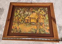 Front view of framed painting showing image of a thatched hut among trees with measuring tape indicating width about 19 inches.