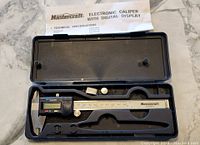 Digital caliper inside black plastic case with foam, LCD display visible, manual included