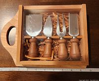 Top-down view of the cheese set inside a wooden box with a transparent lid, showing the four cheese knives with wooden handles arranged neatly in a row.