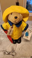 Paddington Bear plush in yellow raincoat and matching hat, holding a red umbrella, standing upright on surface.