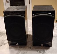 Pair of Precision Acoustics PA 6XB125 speakers standing upright on a marble countertop, front view with fabric grills visible. Both speakers black with wood grain finish.