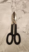 Tin snips lying flat on marble surface, handles black, blade metallic with rust spots visible.